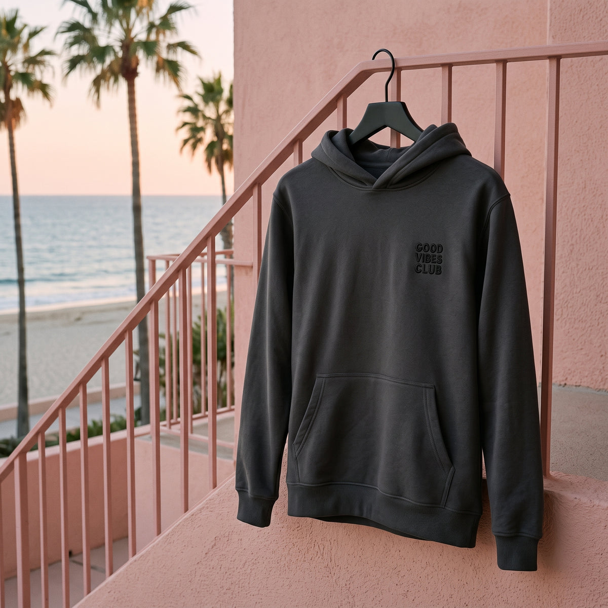 Faded Black Club Hoodie hanging from a pink railing beside the beach.