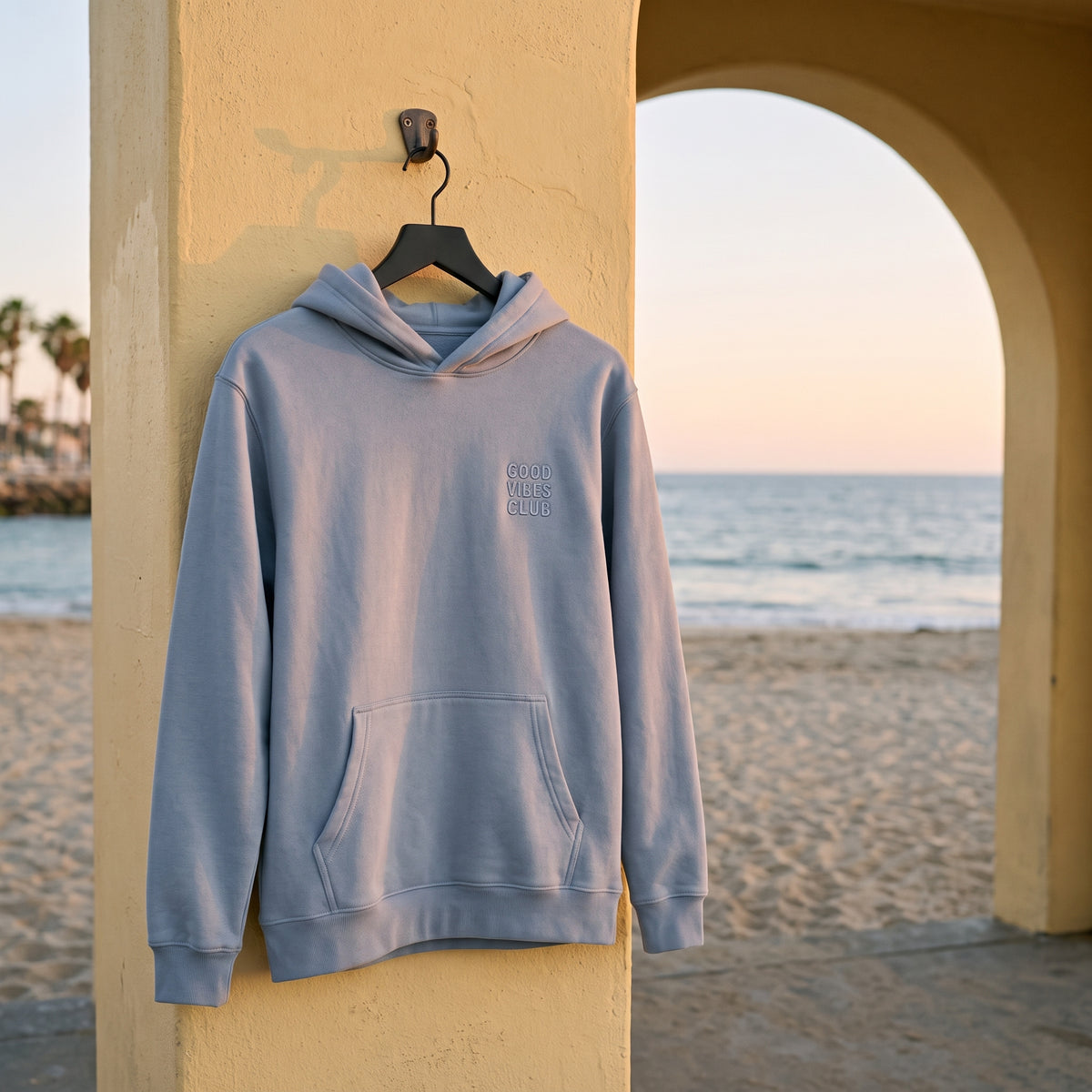 Faded Powder Club Hoodie hanging beside a yellow beachside archway.